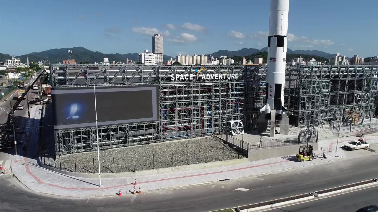 Fachada do Space Adventure, o Museu da NASA em Balneário Camboriú: malha
            estrutural em Light Steel Frame contra o céu, com a réplica do foguetão Saturn V
            ao lado.